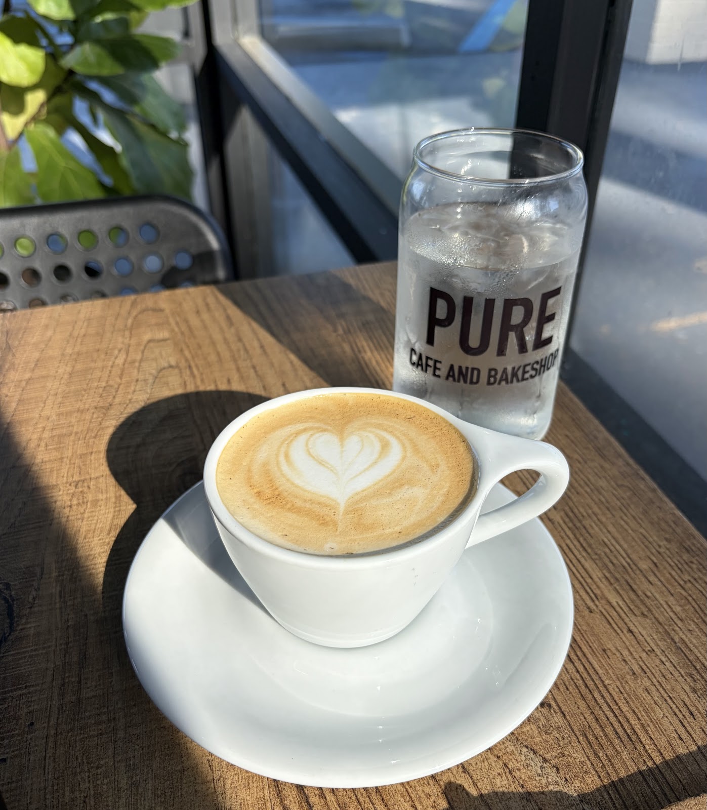 Latte art and a water glass printed with Pure Cafe and Bakeshop on a sunlit table